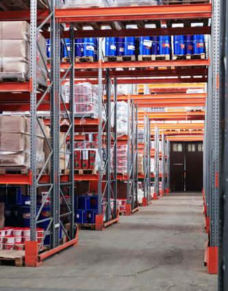 Warehouse aisle with metal shelving stocked with boxes and blue barrels under industrial lighting
