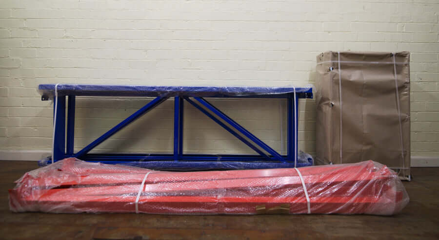 A blue metal frame, red metal bars, and a large flat package wrapped in brown paper are neatly arranged against a light brick wall on a wooden floor.