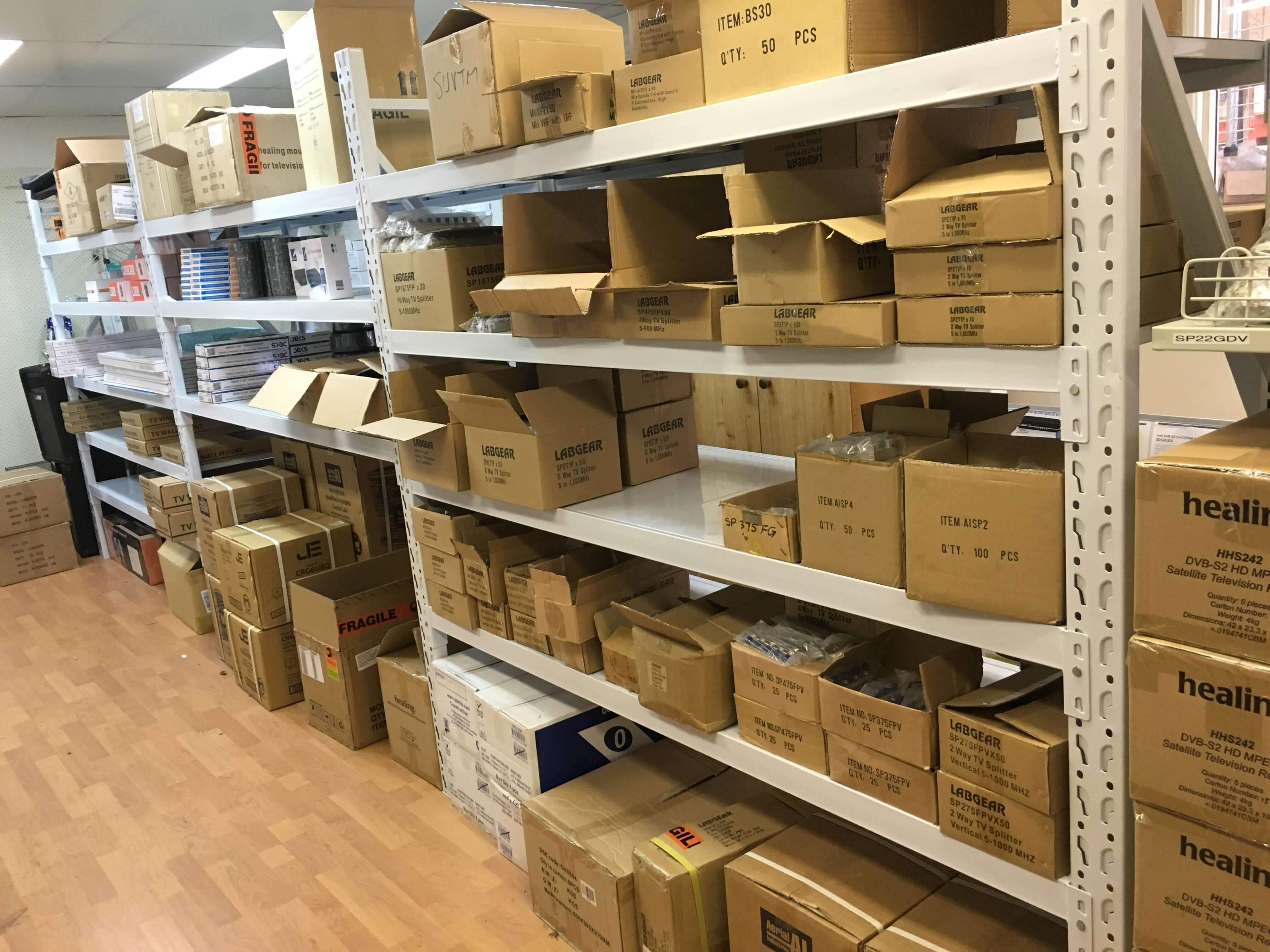 Shelves filled with various cardboard boxes and packages, some labeled, organized along a wall in a storeroom or warehouse with a wooden floor.