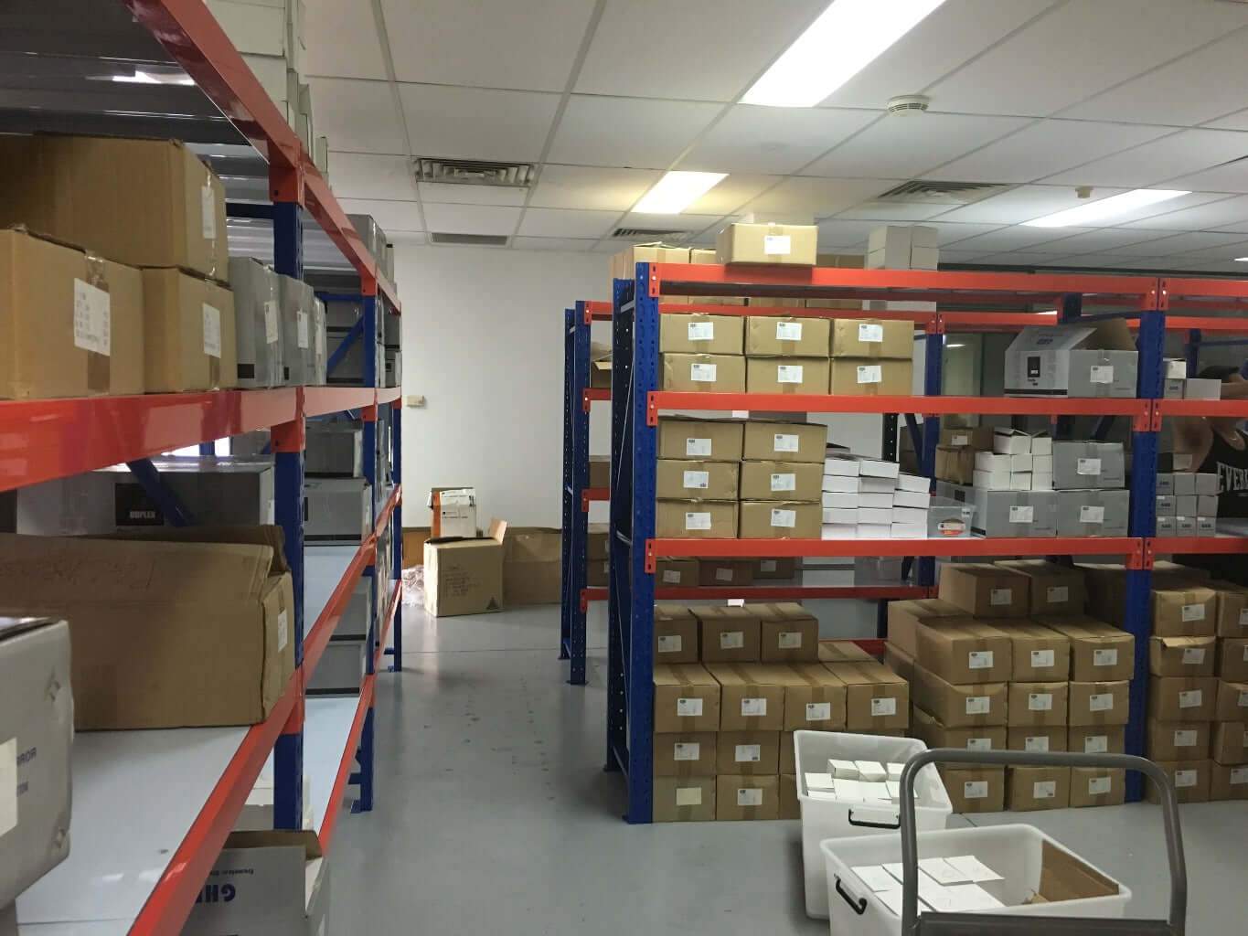 A storage room with blue and orange metal shelves filled with cardboard boxes, labeled and stacked neatly. The floor is gray, and a cart with a white bin is in the foreground. Ceiling lights illuminate the space.